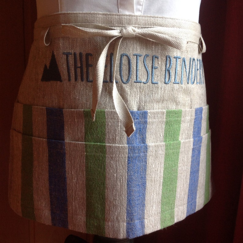 May include: A linen half-apron with a striped pattern in blue and green. The top section features embroidered text that reads "THE MOISE BINDER" above the striped section. The apron has a tie closure and a small pocket.