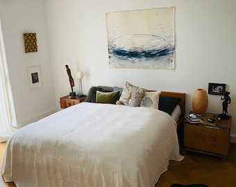 May include: A white bedroom with a large bed with a white linen duvet and a variety of pillows. There is a large abstract painting on the wall above the bed. There is a wooden nightstand with a lamp and a woven basket on the right side of the bed.