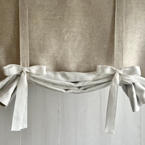 Op de afbeelding: Een paar witte linnen gordijnen met een geplooide, gerimpelde vorm. De gordijnen zijn vastgebonden met bijpassende stofbanden.