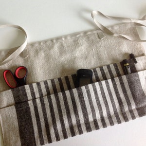 Puede incluir: Delantal de lino beige con un bolsillo a rayas marrones y blancas. El delantal tiene unas tijeras con mangos rojos, un tel&eacute;fono negro y dos bol&iacute;grafos en los bolsillos. Tiene una corbata beige. Ideal para la cocina.