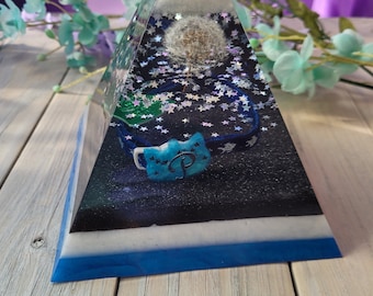 May include: A clear pyramid-shaped resin art piece. Inside, a dandelion seed head is suspended above a blue and black starry background. A blue ribbon with a star-shaped charm is also visible. The base of the pyramid has layers of blue, white, and black.