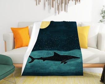 May include: A navy blue throw blanket featuring a whale shark design. The blanket depicts a whale shark swimming in teal water under a starry night sky with a yellow moon. The blanket is draped over a white couch with orange and yellow pillows.