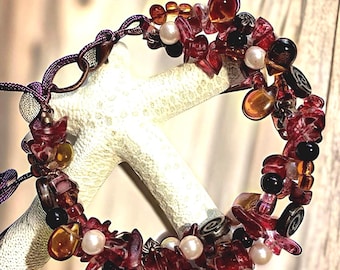 May include: A handcrafted bracelet featuring a mix of red, amber, black, and pearl-colored beads. The beads are irregularly shaped and strung together with a purple cord. The bracelet is displayed on a white, textured surface.