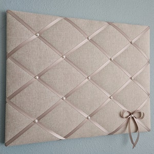 May include: A beige fabric bulletin board with a diamond pattern of white ribbon. The ribbon is held in place with pearl-like tacks. A white bow is attached to the bottom right corner.