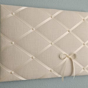 May include: Cream-colored memo board with a linen texture, crisscrossed with ivory ribbons and adorned with faux pearl accents. A decorative bow is tied at the bottom. Ideal for displaying notes or photos.
