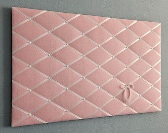 May include: A pink fabric bulletin board with white ribbon crisscrossing the surface. The board is decorated with silver-colored tacks and a white ribbon bow.