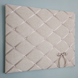 May include: A beige fabric bulletin board with a grid pattern of silver ribbon and silver tacks. A small bow of silver ribbon is attached to the bottom right corner.