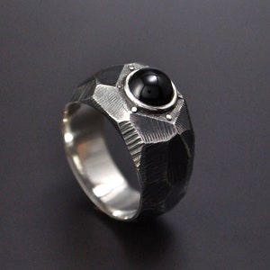 May include: A silver ring with a black gemstone set in a textured, geometric design.