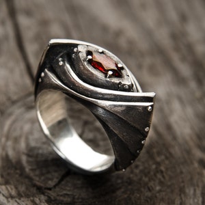 May include: A silver ring with a red gemstone set in a unique, abstract design. The ring has a textured surface and a wide band.