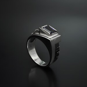 K&ouml;nnte beinhalten: Ein silberner Ring mit einem dunkelvioletten Edelstein, der in einem geometrischen Design gefasst ist. Die Band ist mit kleinen Punkten strukturiert.