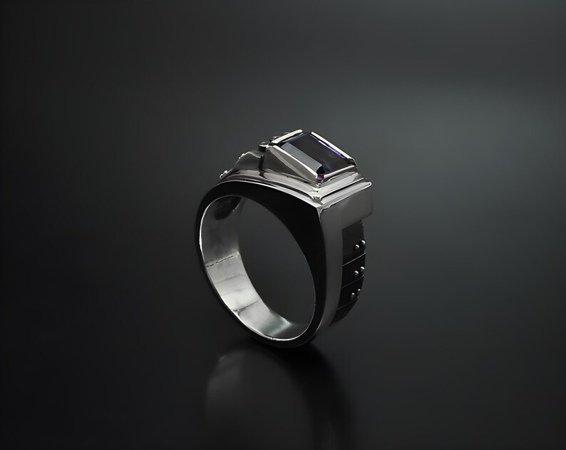 K&ouml;nnte beinhalten: Ein silberner Ring mit einem dunkelvioletten Edelstein, der in einem geometrischen Design gefasst ist. Die Band ist mit kleinen Punkten strukturiert.