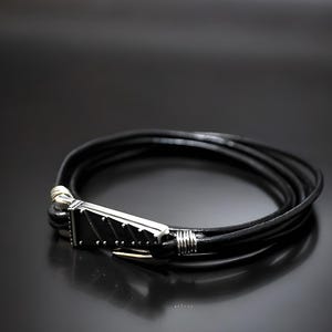 Pu&ograve; includere: Un braccialetto in pelle nera con una chiusura argentata. La chiusura ha un design testurizzato.