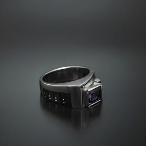 K&ouml;nnte beinhalten: Ein silberner Ring mit einem schwarzen Band und einem violetten Edelstein. Das Band hat ein strukturiertes Design.
