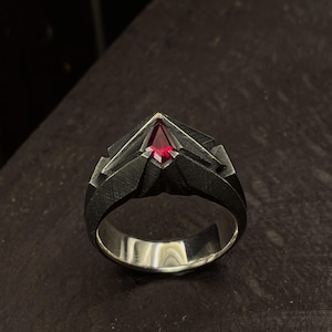 May include: A silver ring with a dark, textured band and a triangular, deep red gemstone. The ring's design features geometric shapes and a polished interior, creating a striking contrast between the dark exterior and the bright gemstone.