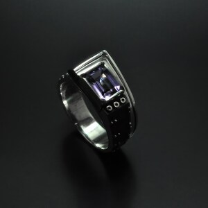 K&ouml;nnte beinhalten: Ein silberner Ring mit einem schwarzen Band und einem violetten Edelstein. Das Band hat kleine silberne Punkte und drei silberne Kreise.
