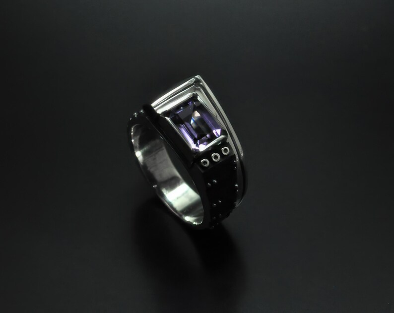 K&ouml;nnte beinhalten: Ein silberner Ring mit einem schwarzen Band und einem violetten Edelstein. Das Band hat kleine silberne Punkte und drei silberne Kreise.