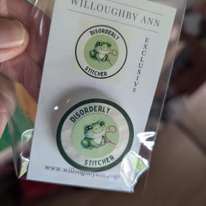 Op de afbeelding: Een ronde pin met een illustratie van een groene kikker en de tekst DISORDERLY STITCHER, verpakt in een doorzichtige plastic hoes met een kaartje waarop WILLOUGHBY ANN, EXCLUSIVE en het websiteadres www.willoughbyann.com staan.
