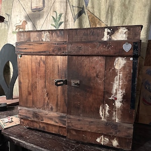 May include: A wooden cabinet with two doors, painted brown. The cabinet has a heart-shaped metal tag on the right door. The cabinet is sitting on a wooden table.