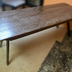 May include: A rustic, rectangular wooden table with a dark brown finish. The table has four slender, dark legs and a distressed appearance, suggesting age and use. The tabletop is approximately 60 inches long.
