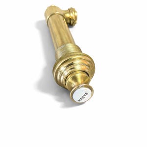May include: A brass-colored plumbing fixture with a cylindrical body and a curved top. The fixture has a white button with the word "WASTE" in black. The object is on a white background.