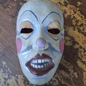 May include: A hand-painted clown mask with a pale grey base, featuring blue arched eyebrows, pink circular cheeks, a small pink nose, and a wide open mouth with dark red lips and visible white teeth. The mask has two oval eye cutouts.