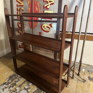 May include: A dark brown wooden shelf unit with four shelves. The shelf unit is standing on a wooden floor with a worn finish.