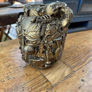 May include: A gold-colored metal vase or planter made from recycled materials. The vase is decorated with a variety of metal objects, including screws, nuts, and bolts.