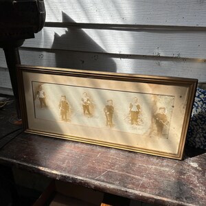 May include: A framed sepia-toned print featuring six figures in vintage attire. The figures are arranged in a row, each holding an object. The frame is a simple gold color, and the print appears aged, suggesting it is an antique.
