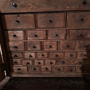 May include: A wooden cabinet with 27 small drawers, each with a black knob and a number from 1 to 27 written on the front. The drawers are arranged in a grid pattern.