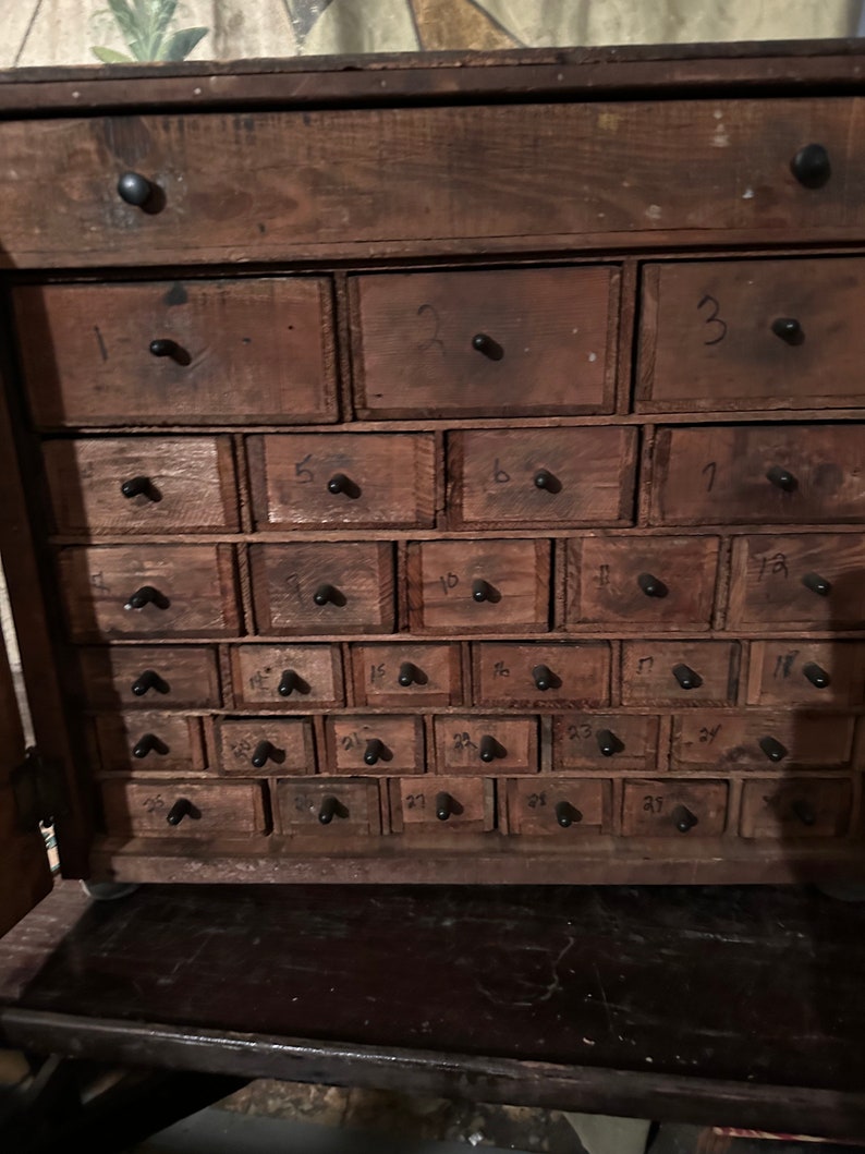 May include: A wooden cabinet with 27 small drawers, each with a black knob and a number from 1 to 27 written on the front. The drawers are arranged in a grid pattern.