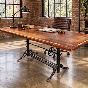 May include: A rectangular wooden desk with a distressed brown finish and a black metal base featuring an adjustable hand crank mechanism. The desk surface is flat and wide, supported by a sturdy industrial-style frame with four splayed feet.