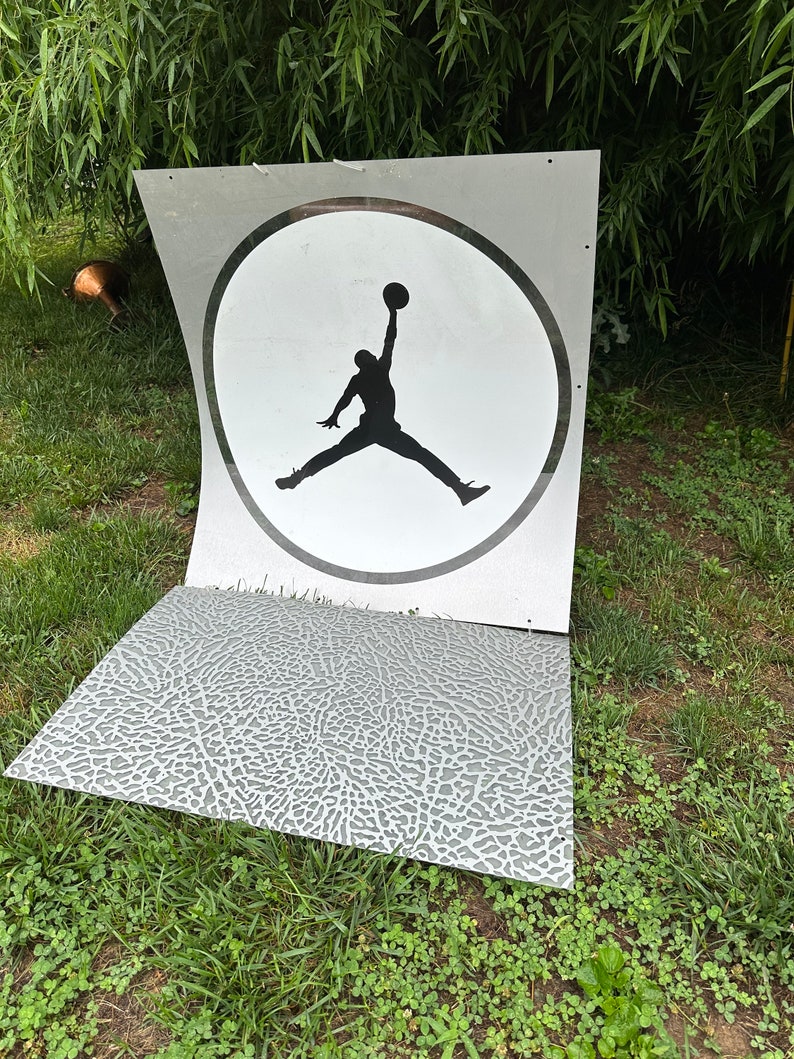 May include: A white and gray sign with a black silhouette of a basketball player jumping in a circle. The sign is on a grassy surface.