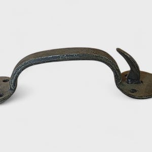 May include: A dark gray, wrought iron handle with an arched grip and decorative, stylized floral end pieces. The handle is designed for a door or gate, with a rustic, antique appearance. The metal has a textured finish.