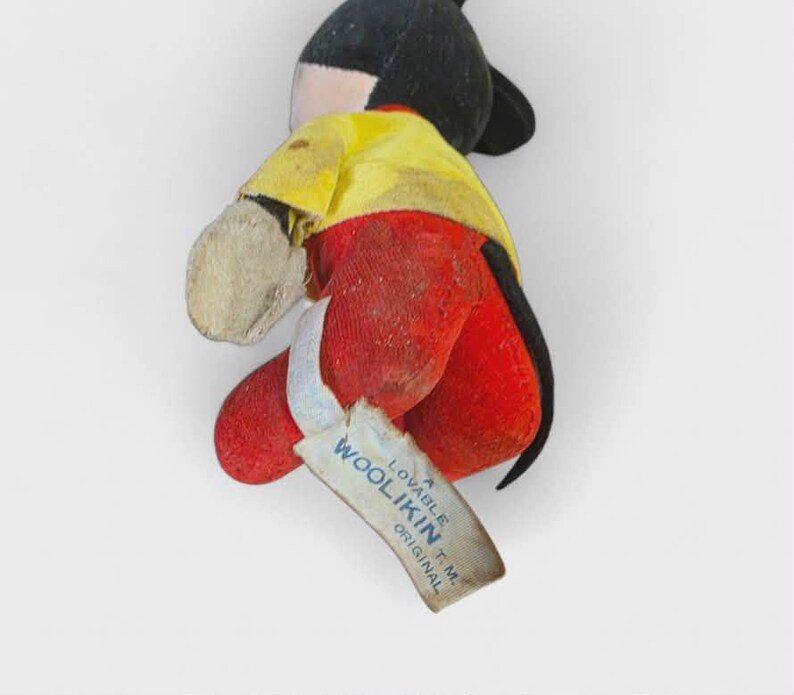 K&ouml;nnte beinhalten: Vintage Mickey Mouse Pl&uuml;schtier. Das Spielzeug hat einen schwarzen Kopf und Ohren, ein gelbes Hemd, rote Shorts und beige Handschuhe. Ein Etikett lautet "Woolikin Original". Das Pl&uuml;schtier ist in einem gut erhaltenen Zustand.