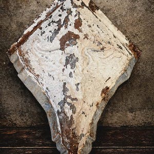 May include: A weathered, diamond-shaped architectural element with peeling white paint, revealing rusty brown and gray layers beneath. The piece has a textured surface and is set against a dark wooden background.