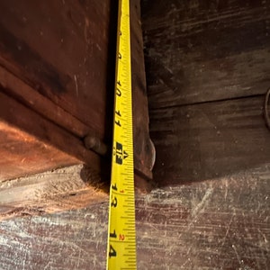 May include: Close-up of a yellow measuring tape with black markings, showing measurements from 10 to 15 inches. The tape is positioned against a dark brown wooden surface.