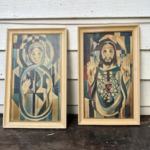 May include: Two rectangular framed wall art pieces featuring geometric religious portraits. The left print depicts a figure in blue and gold tones with a halo. The right print shows a bearded figure with raised hands and a central heart emblem.