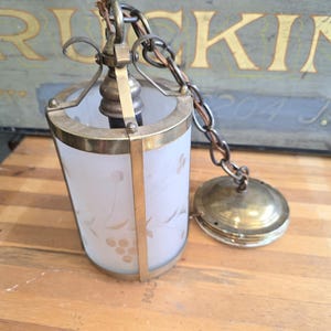 May include: Vintage brass and frosted glass pendant light fixture. The cylindrical shade features a delicate floral design. The fixture includes a chain and a decorative ceiling cap. The metal components have a warm, aged patina.