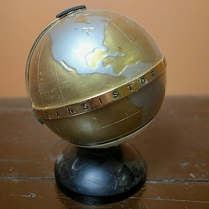 May include: A white ceramic mug with a black handle and the text "But First, Coffee" in a bold, black font. The mug is set against a blurred background, suggesting a cozy setting.