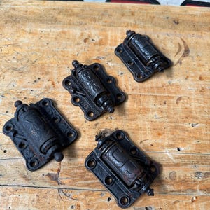 May include: Four antique black metal hinges with decorative detailing. Each hinge has a rectangular base with rounded corners and multiple screw holes. The central barrel of each hinge is cylindrical. The hinges are arranged on a weathered wooden surface.