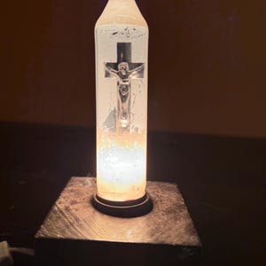 May include: A religious-themed lamp featuring a clear, cylindrical glass shade with a silver crucifix. The lamp emits a warm, soft glow, and is mounted on a dark, square base. The overall design is simple and elegant.