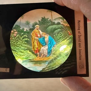 May include: A vintage slide featuring a colorful illustration of two figures in a garden setting. One figure, in a gold and red robe, is embracing another in a blue robe. The text "Meeting of David and Jonathan" is visible.