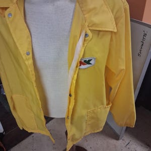 May include: A bright yellow windbreaker jacket featuring a pointed collar, long sleeves, and silver-toned snap button closures. A white patch with the red and green letters AK is stitched on the left chest. The jacket is worn over a white sweater.
