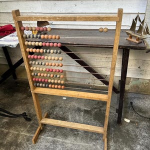 May include: A vintage wooden abacus with multiple rows of colorful beads. The frame is made of light-colored wood, and the beads are in shades of red, orange, yellow, and gray. The abacus is standing on a concrete surface.