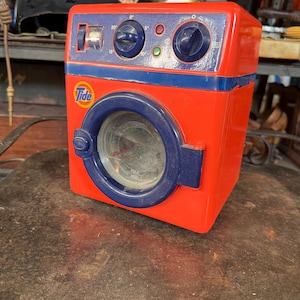 May include: A vintage toy washing machine in red and blue. The toy has a round door, dials, and the Tide logo. The toy is sitting on a dark metal surface.