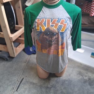May include: A gray and green baseball-style shirt with three-quarter length sleeves. The front features a graphic of a skull wearing sunglasses in front of a pyramid, with the text KISS above and 1988 below the skull in orange and yellow lettering.