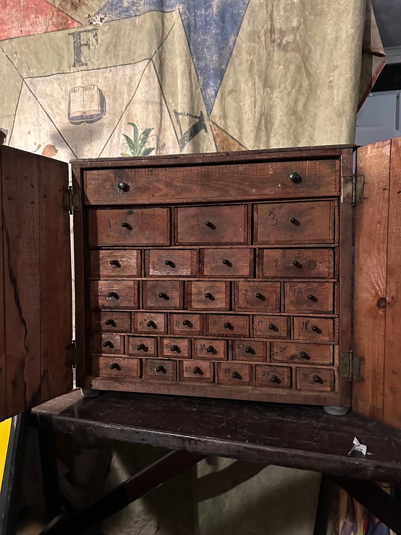 May include: A wooden cabinet with 30 small drawers, each with a black knob. The drawers are numbered 1 through 30. The cabinet is open and the drawers are visible. The cabinet is made of dark wood and has a rustic appearance.