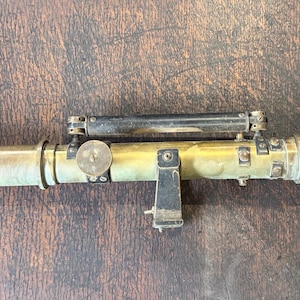 May include: A vintage brass telescope featuring a cylindrical body with a metallic finish, a black mounting bracket, and a secondary black tubular component attached to the top. The instrument displays visible signs of age and oxidation on the metal.
