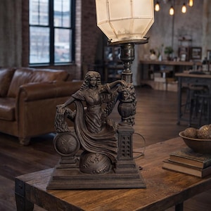May include: A bronze-toned table lamp with a geometric shade. The lamp features a sculpted base with a seated female figure, a globe, and decorative elements. The lamp is placed on a wooden table.