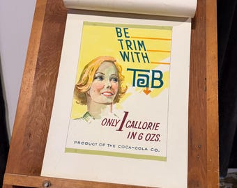 May include: Vintage advertisement for Tab soda, displayed in a wooden frame. The poster features a watercolor illustration of a woman and the text "BE TRIM WITH Tab." It also states "ONLY 1 CALLORIE IN 6 OZS." The product is from the Coca-Cola Co.